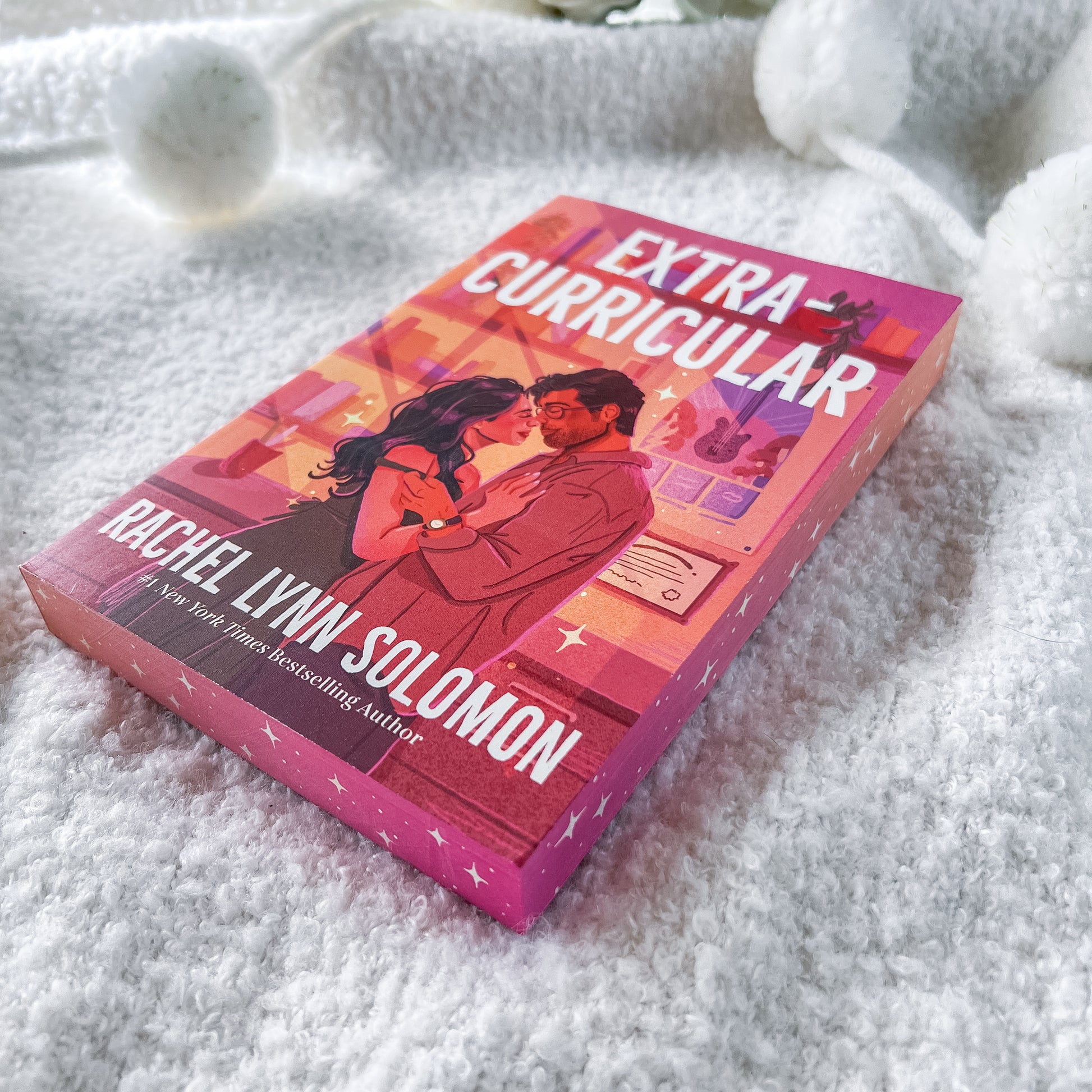 A boxed book titled "Extra Curricular" by Rachel Lynn Solomon lies on a soft white textured blanket. The cover art shows a couple embracing in a warm pink and orange-toned room, with the title and author name in large white text.