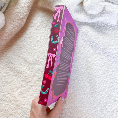 A hand holds a hardcover book titled 'The Horse Whisperer' by Jason Reynolds. The book's cover features a vibrant purple and pink design with illustrations of pink bows, red cherries, and turquoise horseshoes.