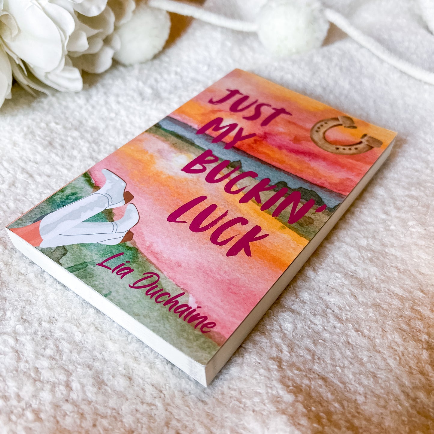 A book titled 'Just My Bucking Luck' by Lia Duchaine, featuring a colorful watercolor cover with pink, orange, and green tones, resting on a soft white textured blanket with white flowers in the background.