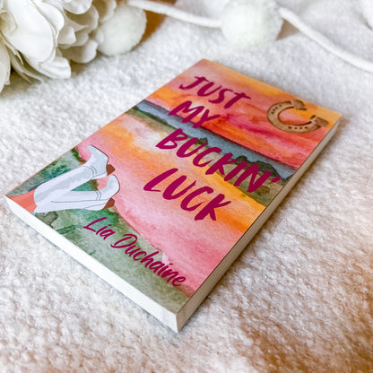 A book titled 'Just My Bucking Luck' by Lia Duchaine, featuring a colorful watercolor cover with pink, orange, and green tones, resting on a soft white textured blanket with white flowers in the background.