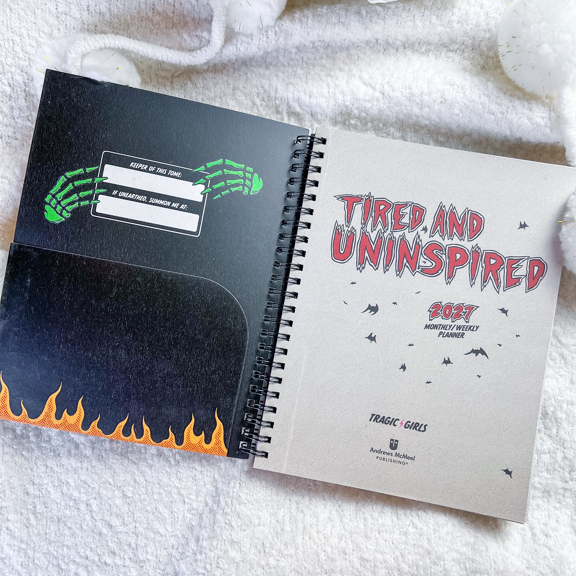 An open spiral-bound planner or notebook lying on a white textured blanket. The left page is black with a green skeleton hand graphic, a date box, and orange flames along the bottom.