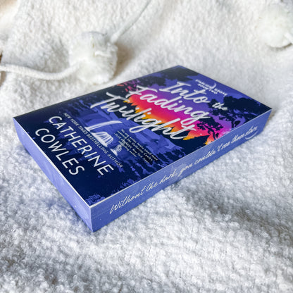 the book 'Into the Fading Twilight' by Catherine Cowles, placed on a soft white textured blanket. The box features a dark blue and purple cover with a sunset graphic and white title text.