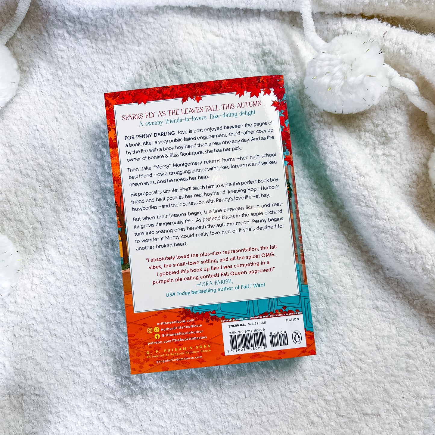 The back cover of the paperback book photographed flat on a white textured blanket. The cover features a vibrant red and orange fiery design on the top half and a teal blue section on the bottom right containing the barcode and author details.