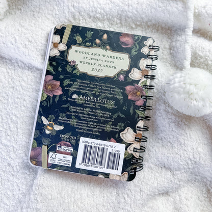 A 2027 weekly planner notebook titled 'Woodland Wardens' by Jessica Bour, published by Amber Lotus, shown from its back cover. The cover features a dark navy blue background decorated with illustrations of woodland flowers (pink, white, and green) and a bee.
