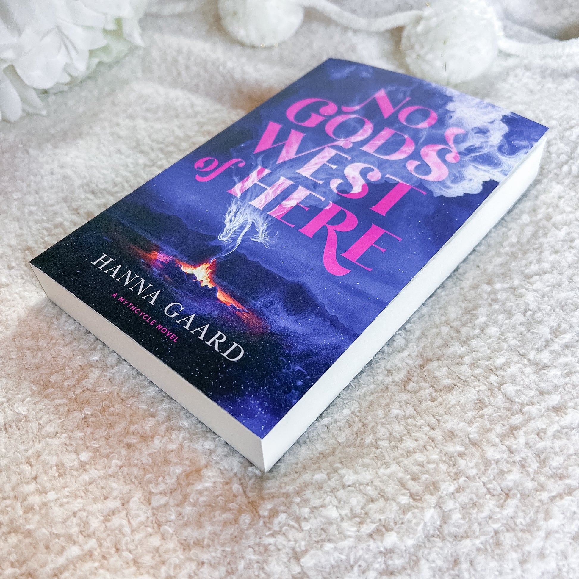 A  book titled 'No Gods or Here' by Hanna Gaard, lying on a textured cream-colored blanket with soft white decorative flowers in the background. The book cover features a dark blue and purple cosmic illustration with pink title text.