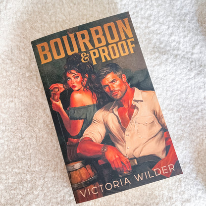 A paperback book titled 'Bourbon & Proof' by Victoria Wilder, featuring a vintage-style illustrated cover of a man and woman in a warm, amber-toned scene, photographed on a textured off-white fabric background.