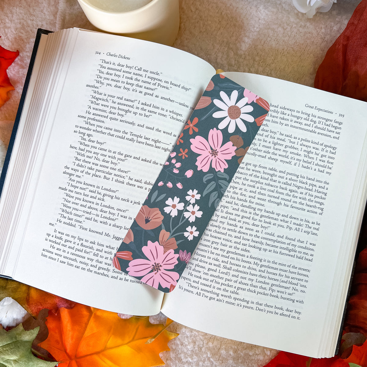 A floral book bookmark with pink, white, and orange flowers on a dark background, placed on an open book surrounded by autumn leaves and a cup.