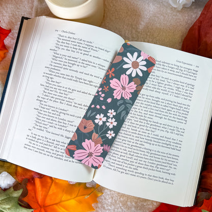 A floral book bookmark with pink, white, and orange flowers on a dark background, placed on an open book surrounded by autumn leaves and a cup.