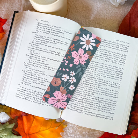A floral book bookmark with pink, white, and orange flowers on a dark background, placed on an open book surrounded by autumn leaves and a cup.