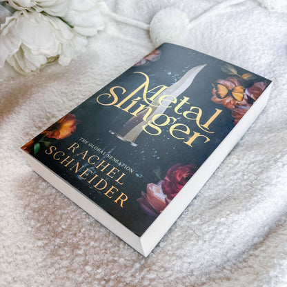 A book titled 'Metal Slinger' by Rachel Schneider lies on a soft white textured blanket, with white flowers in the upper left corner. The book cover is dark with gold lettering and floral illustrations.