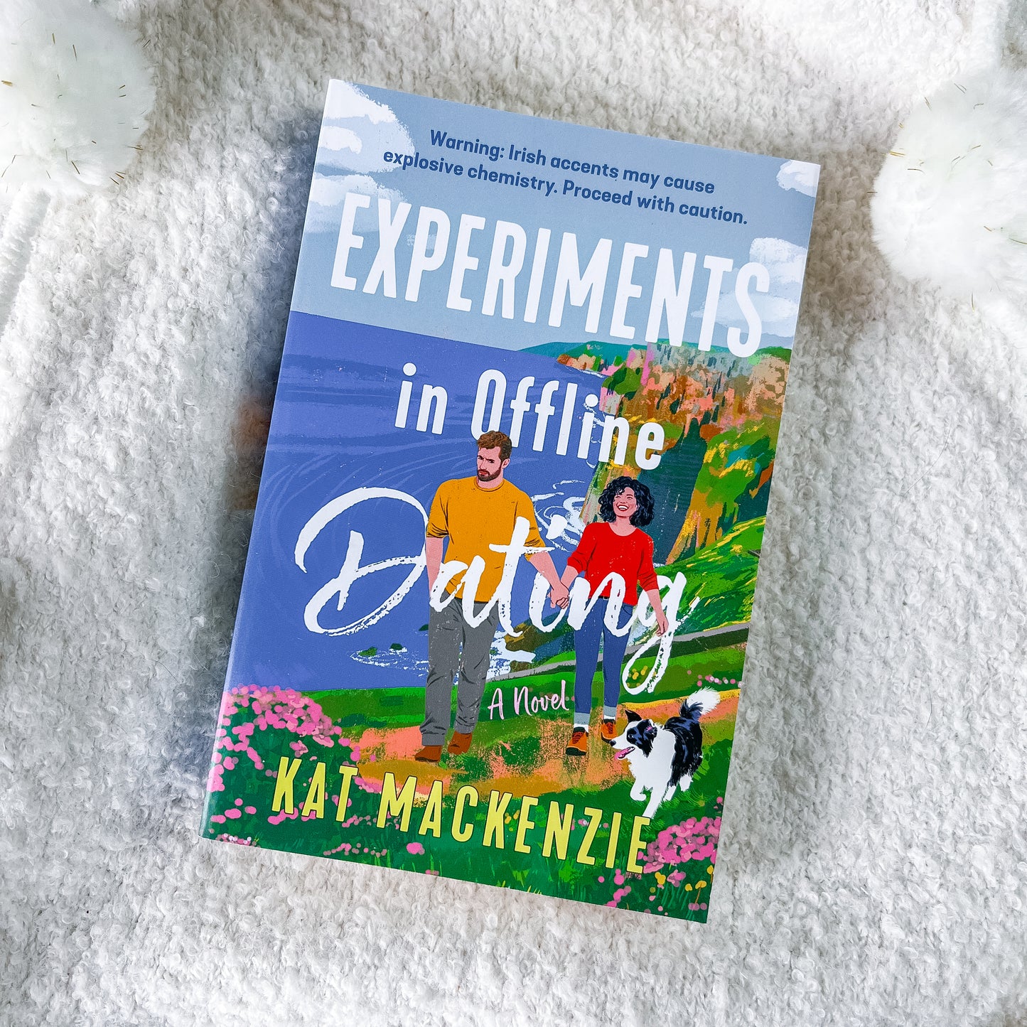 The book cover of 'Experiments in Offline Dating' by Kat Mackenzie, featuring a colorful illustration of a couple and their dog on a hillside, displayed on a white textured blanket.
