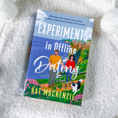 The book cover of 'Experiments in Offline Dating' by Kat Mackenzie, featuring a colorful illustration of a couple and their dog on a hillside, displayed on a white textured blanket.
