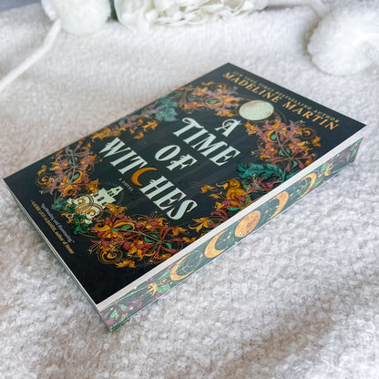 A Time of Witches book by Madeline Martin, featuring a dark cover with intricate gold, orange, and green witch illustrations, resting on a white textured blanket.
