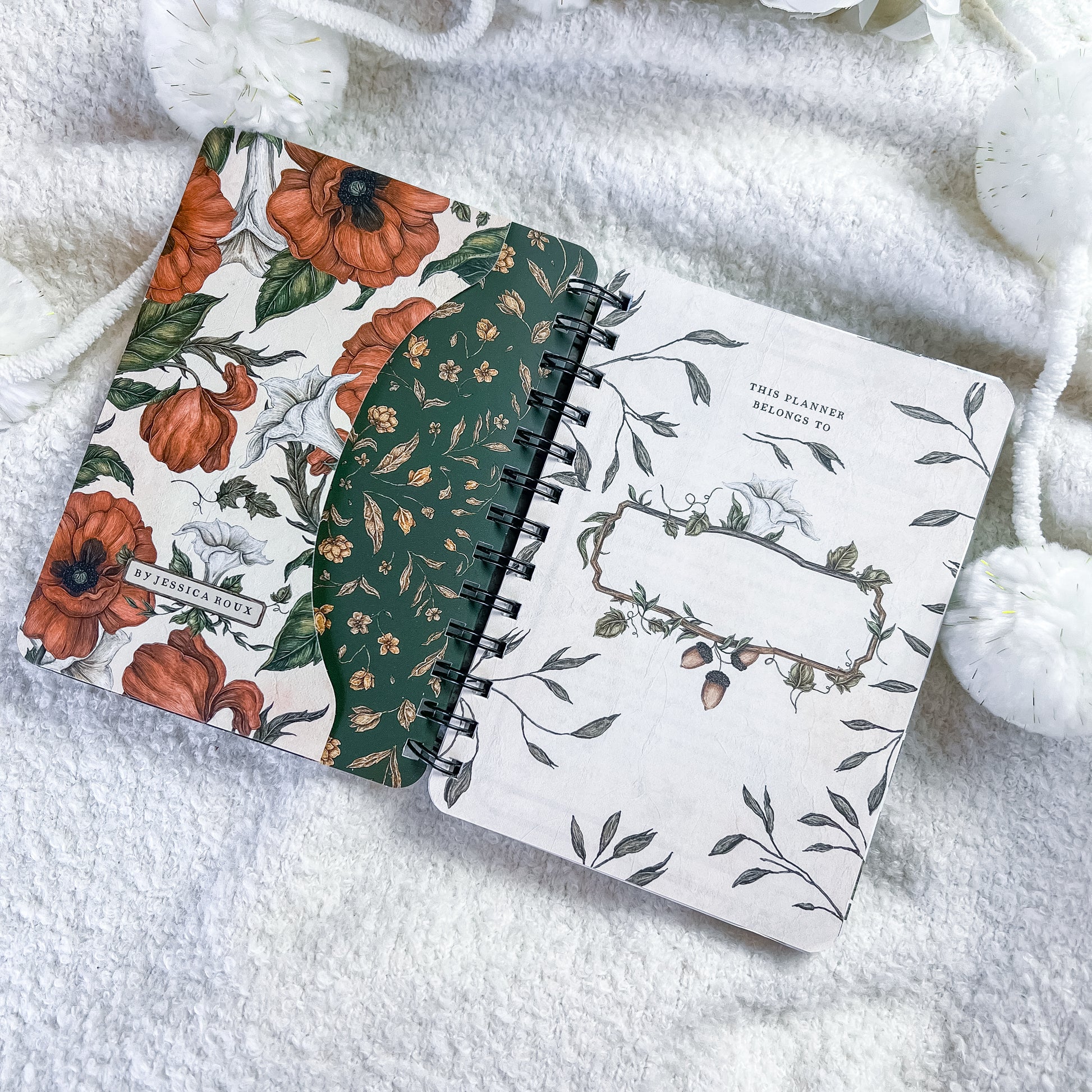 A spiral-bound planner or notebook lies open on a soft white textured blanket, surrounded by white decorative pom-pom balls. The cover has a floral print with orange flowers, green leaves, and a dark green vertical panel with small floral motifs.