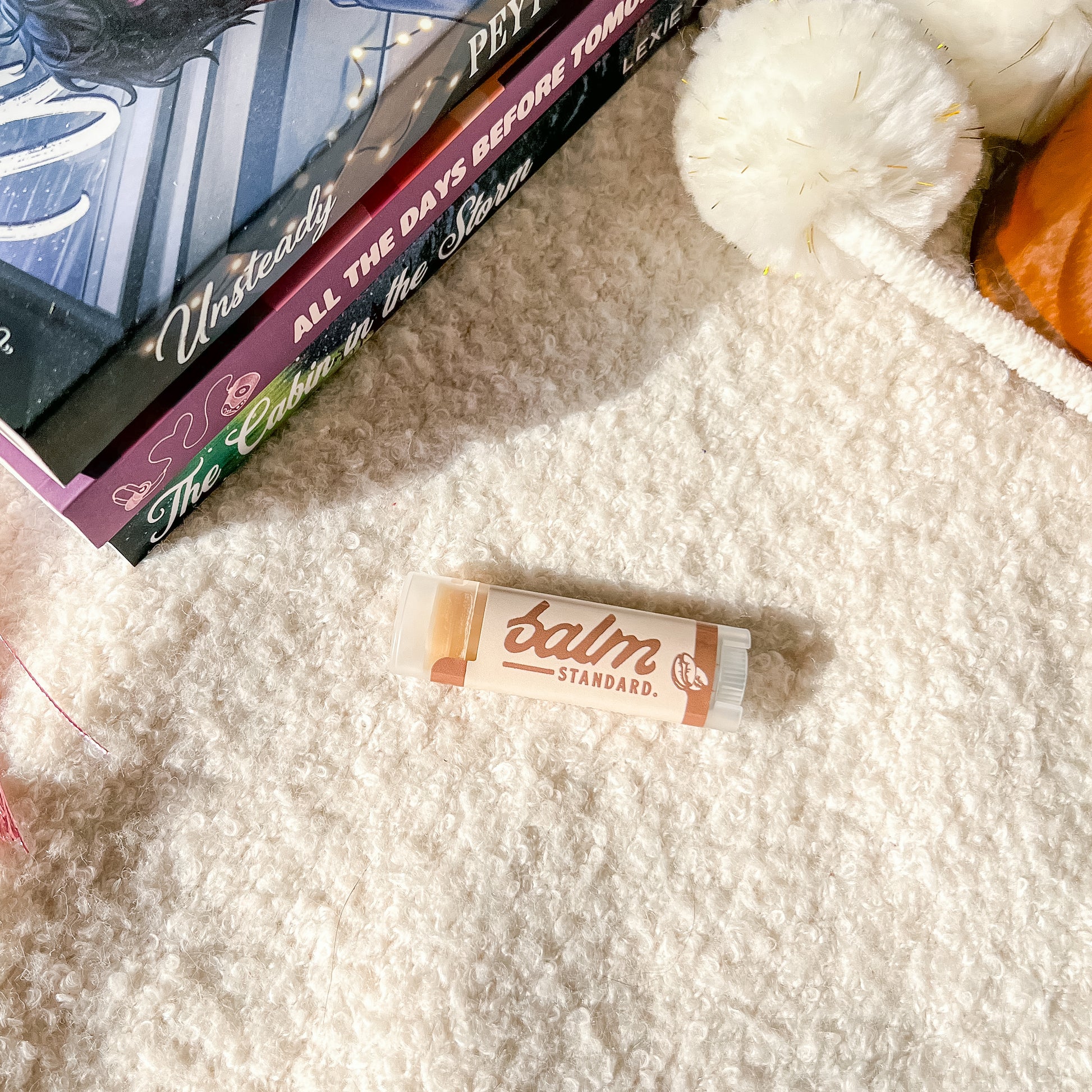 A tube of lip balm labeled 'balm STANDARD.' lies on a cream shag rug next to stacked books and a fluffy white pom-pom toy.