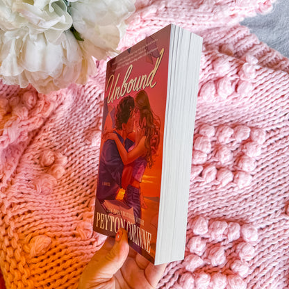Book titled 'Unbound' by Peyton Corinne held against a pink textured fabric background with flowers.