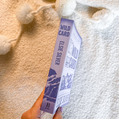 A hand holds a book titled 'Wild Card' by Else Silver, featuring a lavender and purple cover with mountain illustrations, set against a cozy white textured blanket with white pom-pom tassels.