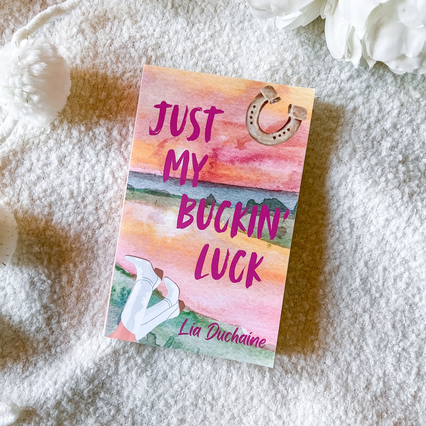A book titled 'Just My Buckin' Luck' by Lia Duchaine lies on a soft white textured blanket, styled with white flowers in the corners. The cover features a watercolor sunset design in pink, orange, and yellow, with a horseshoe graphic and an illustration of white boots.