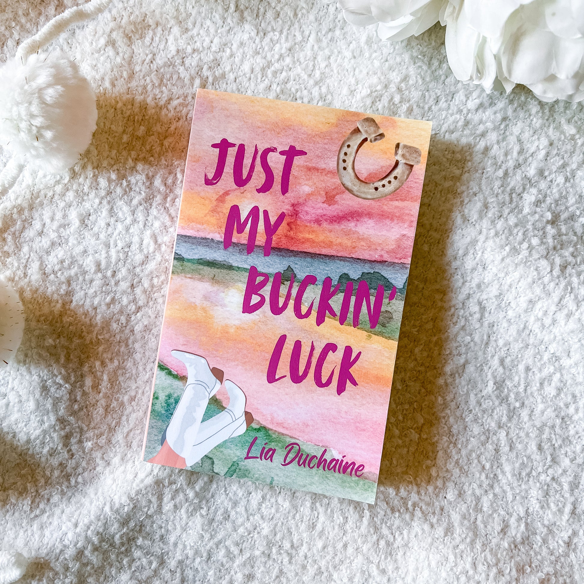 A book titled 'Just My Buckin' Luck' by Lia Duchaine lies on a soft white textured blanket, styled with white flowers in the corners. The cover features a watercolor sunset design in pink, orange, and yellow, with a horseshoe graphic and an illustration of white boots.
