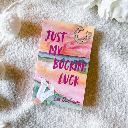 A book titled 'Just My Buckin' Luck' by Lia Duchaine lies on a soft white textured blanket, styled with white flowers in the corners. The cover features a watercolor sunset design in pink, orange, and yellow, with a horseshoe graphic and an illustration of white boots.