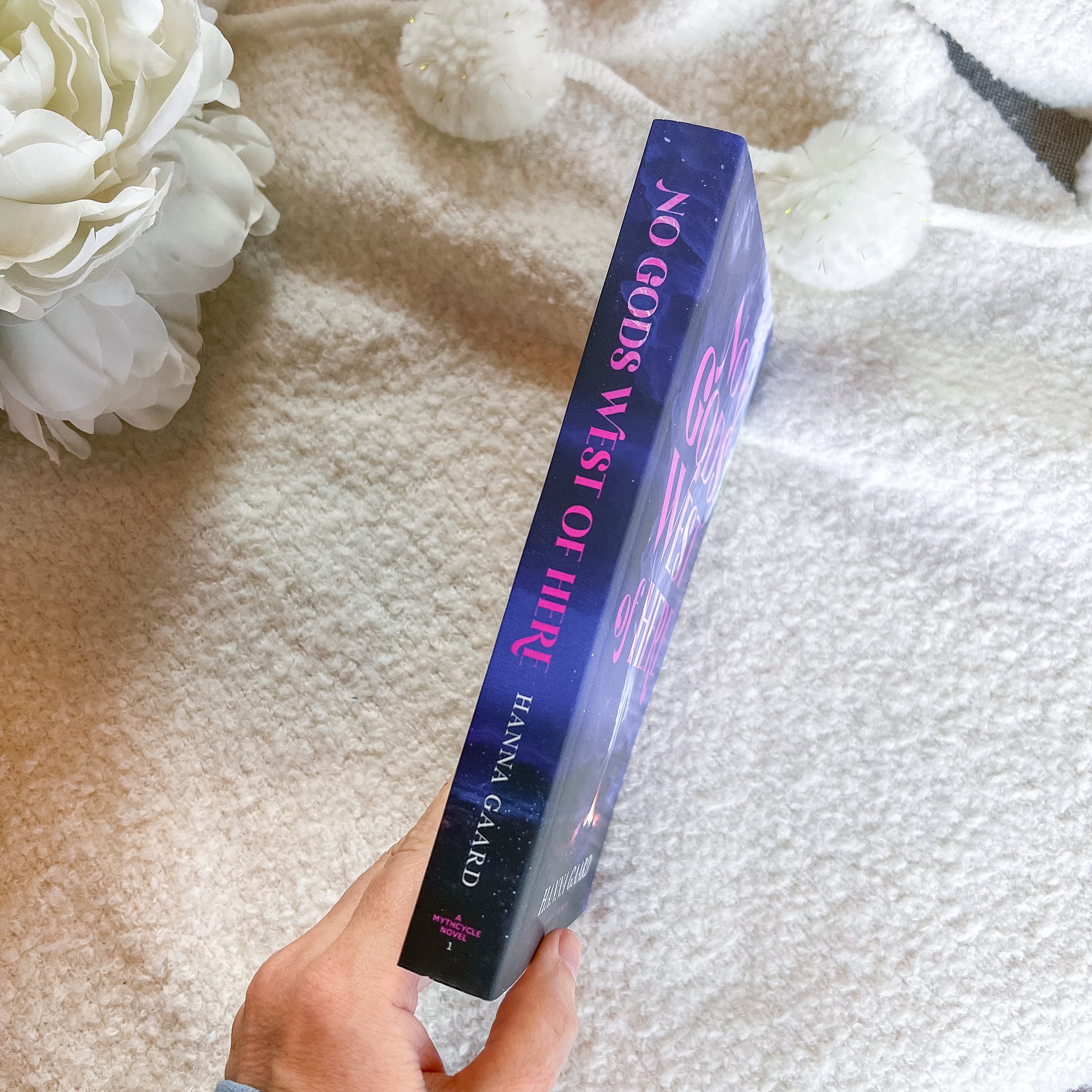 A hand holds a book titled 'No Gods West of Here' , showing the spine with pink lettering on a dark blue textured background, set against a soft cream blanket with white pom-poms and a white flower.