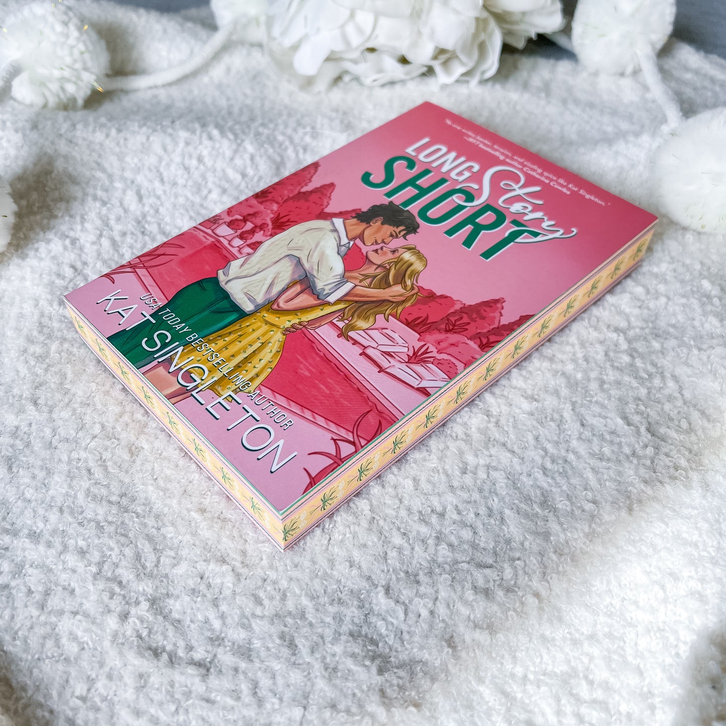 A paperback copy of the book 'Long Story Short' by Kat Singleton lies on a soft white textured blanket with decorative white flowers in the background. The cover features a pink background with an illustration of a couple embracing, with the title in large green and white text.