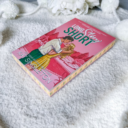 A paperback copy of the book 'Long Story Short' by Kat Singleton lies on a soft white textured blanket with decorative white flowers in the background. The cover features a pink background with an illustration of a couple embracing, with the title in large green and white text.