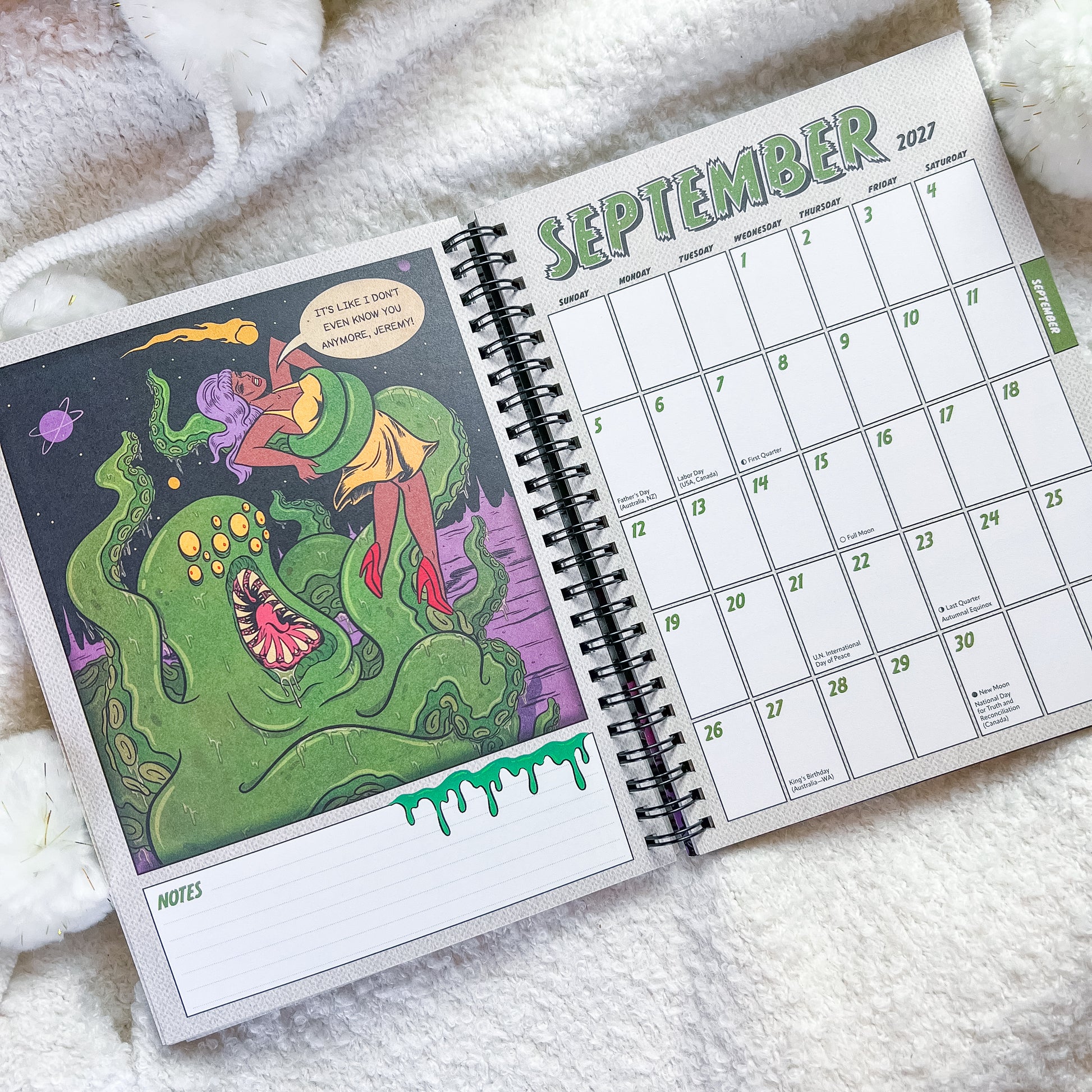 An open spiral-bound planner or calendar showing September 2027, with a colorful illustrated left page and a monthly calendar grid on the right, placed on a soft white textured blanket.
