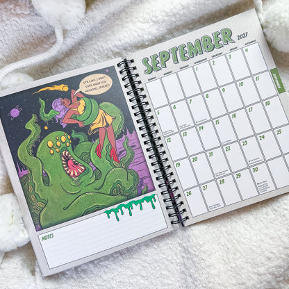 An open spiral-bound planner or calendar showing September 2027, with a colorful illustrated left page and a monthly calendar grid on the right, placed on a soft white textured blanket.
