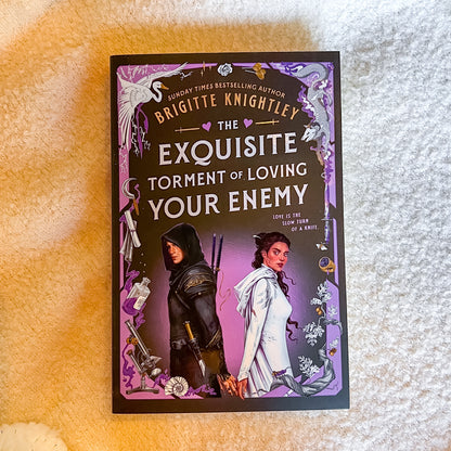 The book cover of 'The Exquisite Torment of Loving Your Enemy' by Brigitte Knightley, featuring a dark purple and black background with fantasy illustrations of two characters and white title text, photographed on a textured cream fabric background.