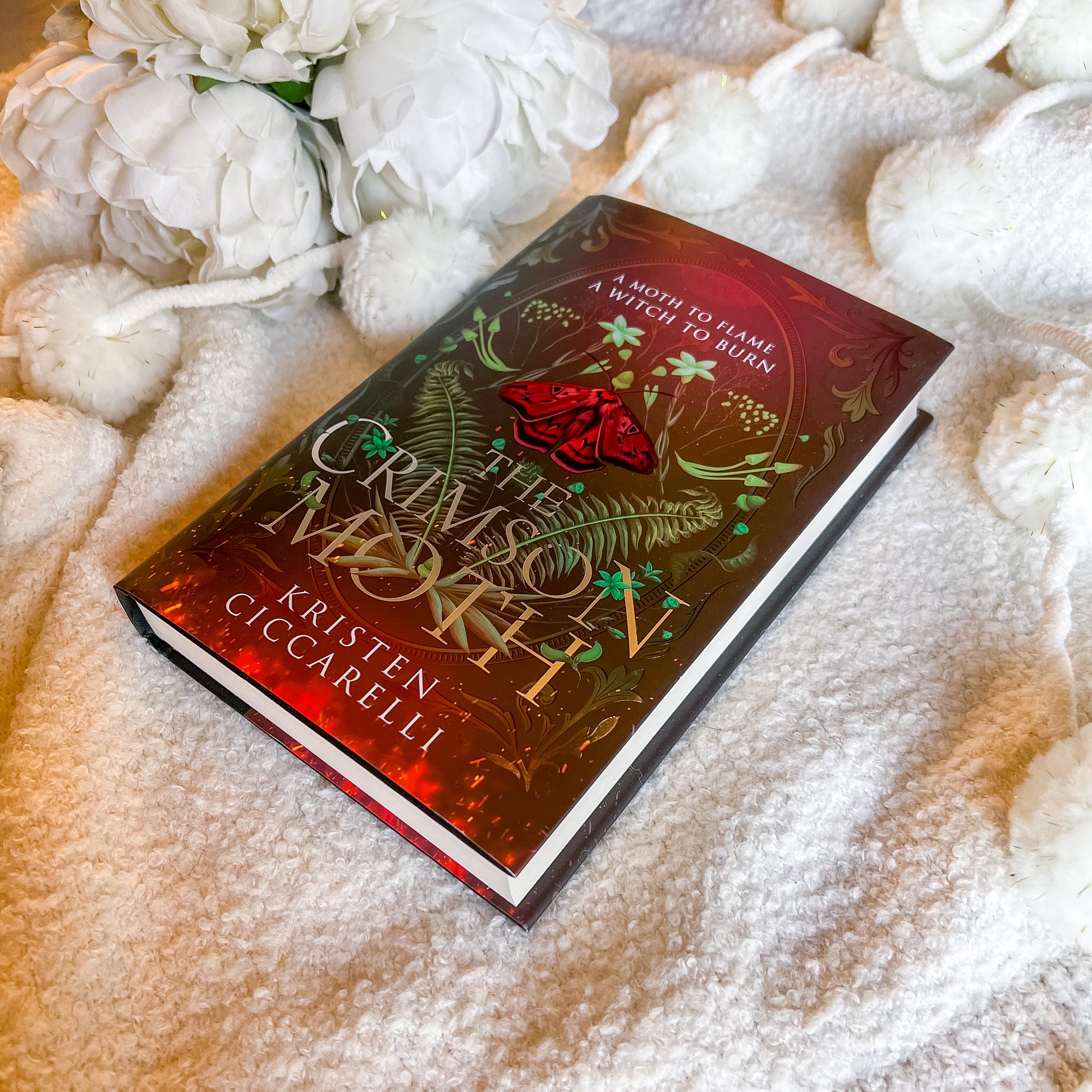 Book titled 'The Crimson Moth' by Kristen Ciccarelli on a textured surface with white flowers.