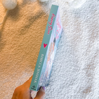A hand holds a copy of the book, showing the teal spine and front cover against a soft beige carpet background.