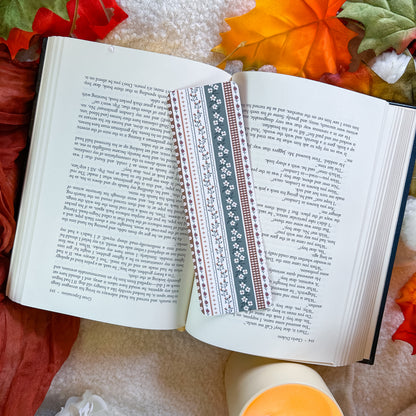 A decorative paper bookmark with a blue and white floral pattern and red and white geometric borders, placed on an open book surrounded by autumn leaves and a candle.