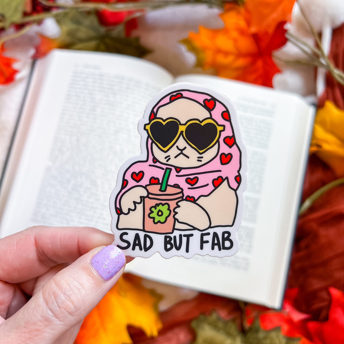 A hand with lavender nail polish holds a die-cut sticker featuring a cartoon cat in a pink heart-patterned hijab, wearing heart-shaped sunglasses and holding a drink. The sticker reads 'SAD BUT FAB' in black text.