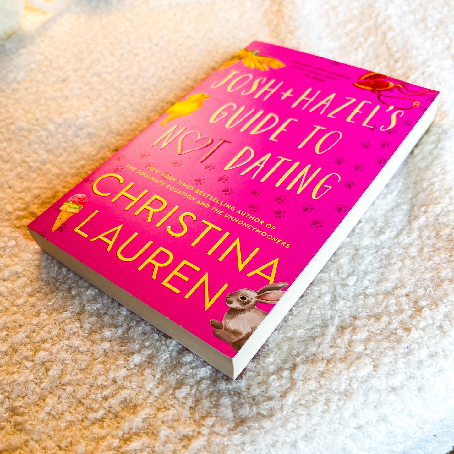 A hardcover book titled 'Josh + Hazels Guide to Not Dating' by Christina Lauren, featuring a bright pink cover with yellow text and illustrations of animals, placed on a textured beige carpet.