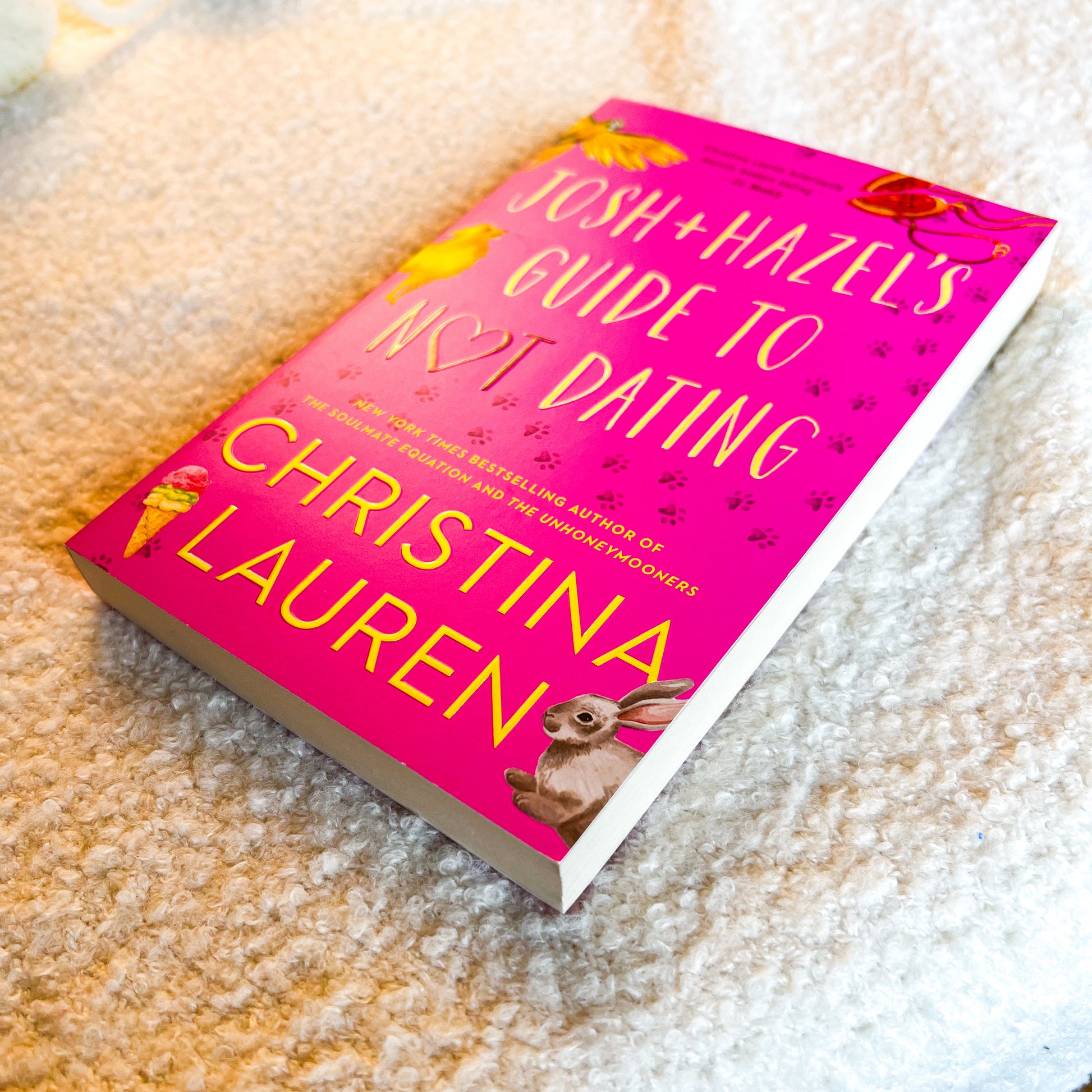 A hardcover book titled 'Josh + Hazels Guide to Not Dating' by Christina Lauren, featuring a bright pink cover with yellow text and illustrations of animals, placed on a textured beige carpet.