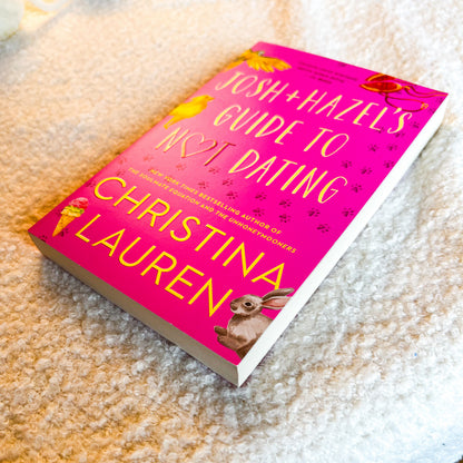 A hardcover book titled 'Josh + Hazels Guide to Not Dating' by Christina Lauren, featuring a bright pink cover with yellow text and illustrations of animals, placed on a textured beige carpet.