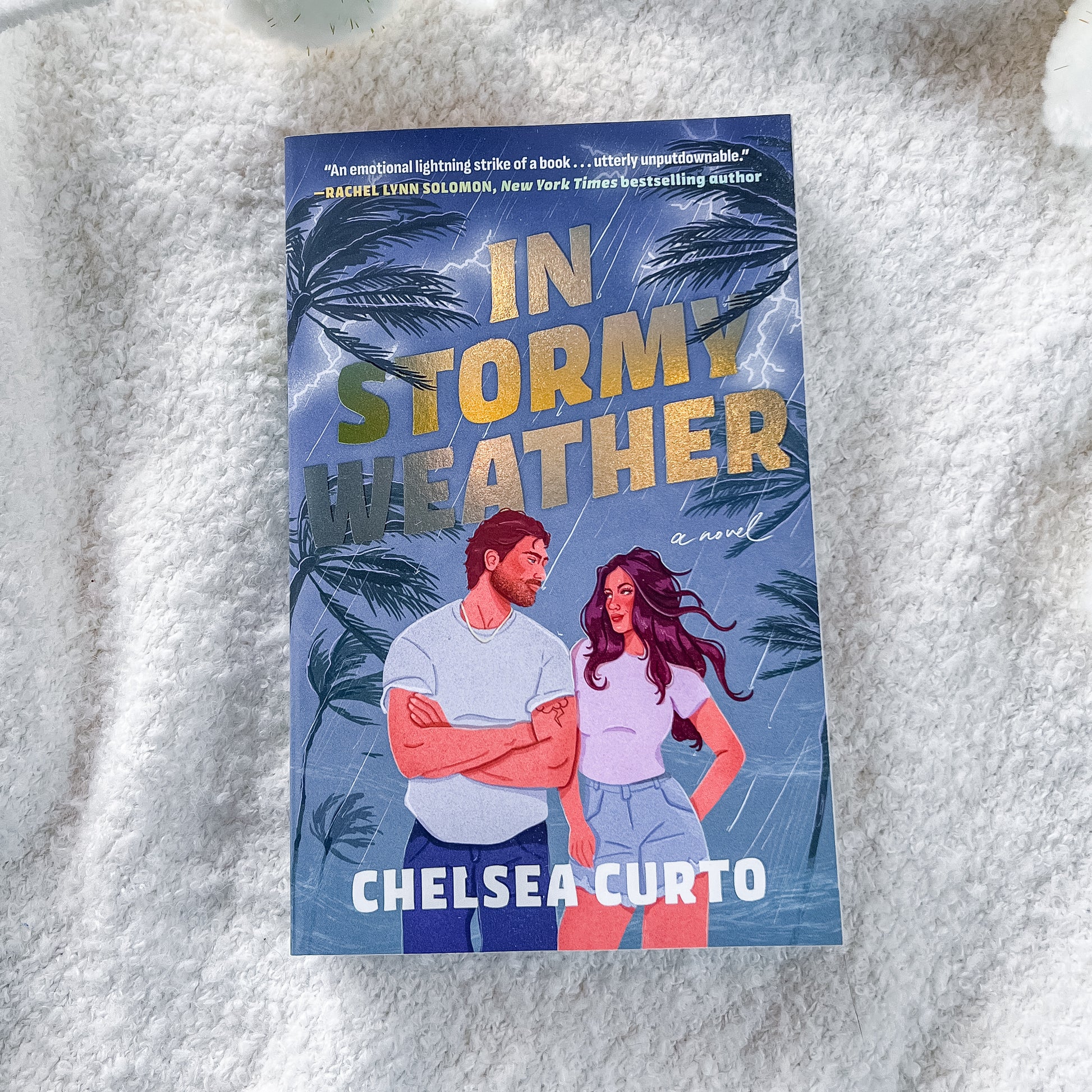 The book cover of 'In Stormy Weather' by Chelsea Curto, featuring a blue tropical storm background with palm trees and an illustration of a man and woman, placed on a white textured blanket.
