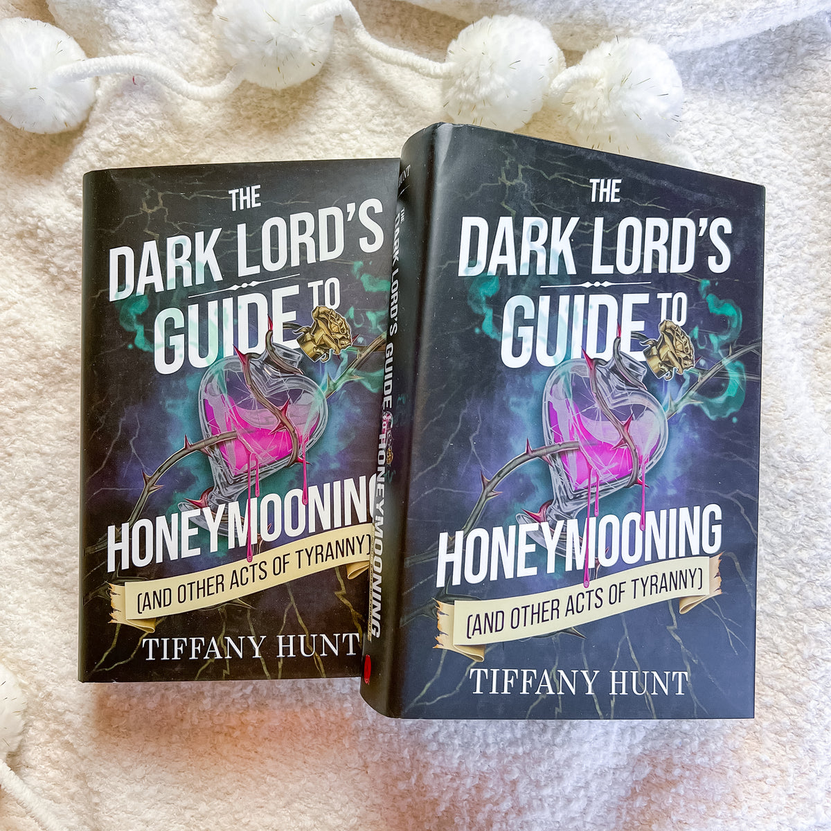 Two copies of the book 'The Dark Lord's Guide to Honeymoonings' by Tiffany Hunt, displayed side-by-side on a soft, textured cream fabric background with white pom-pom beads hanging at the top.