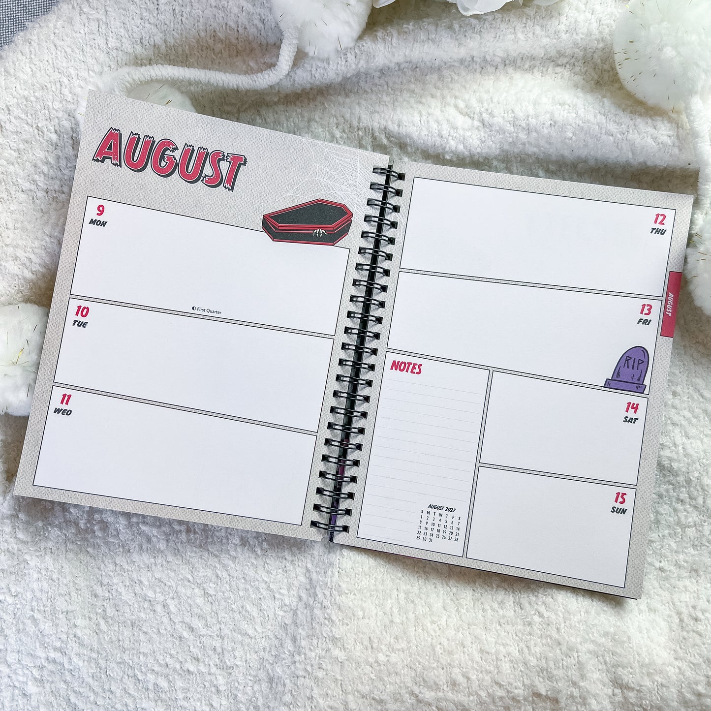 An open spiral-bound planner showing a weekly layout for August, placed on a soft white textured blanket.
