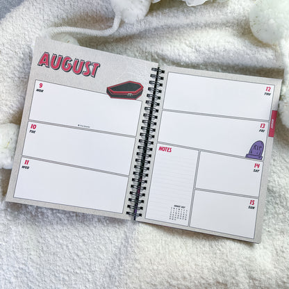 An open spiral-bound planner showing a weekly layout for August, placed on a soft white textured blanket.