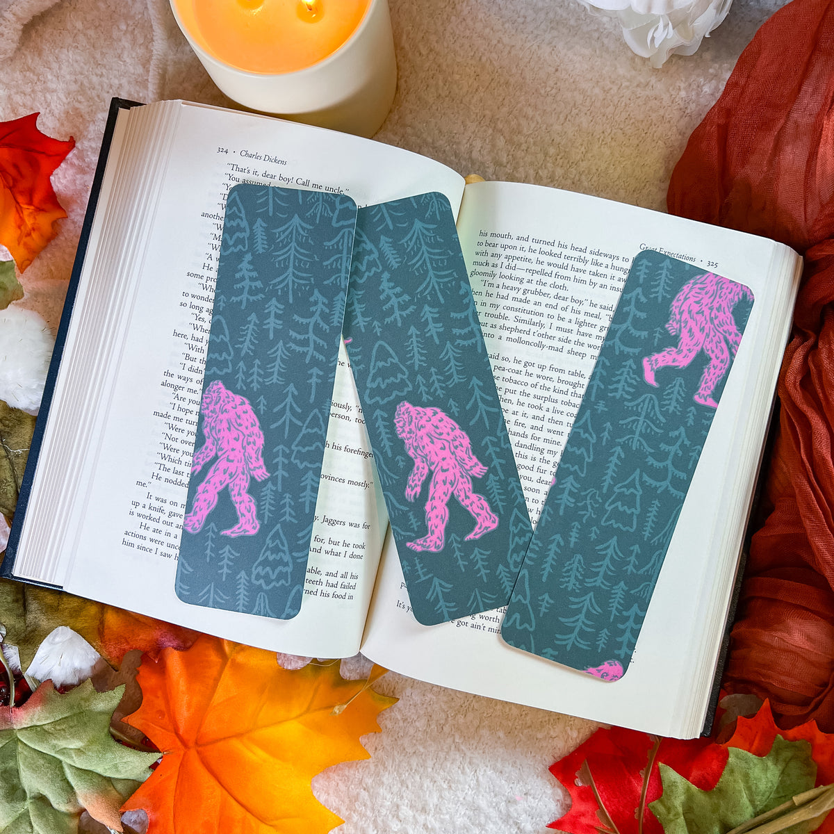 rectangular bookmarks with a teal forest pattern and pink Bigfoot designs are placed on an open book, surrounded by autumn leaves, a candle, and fabric.