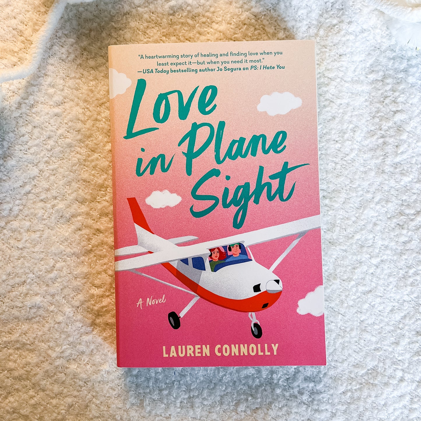 The book cover of 'Love in Plane Sight' by Lauren Connolly, featuring a pink and orange gradient background with a small red and white airplane, placed on a textured beige fabric surface.