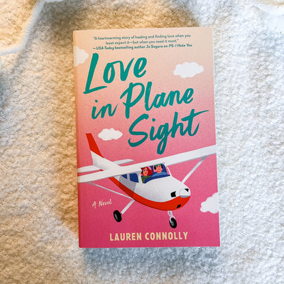 The book cover of 'Love in Plane Sight' by Lauren Connolly, featuring a pink and orange gradient background with a small red and white airplane, placed on a textured beige fabric surface.