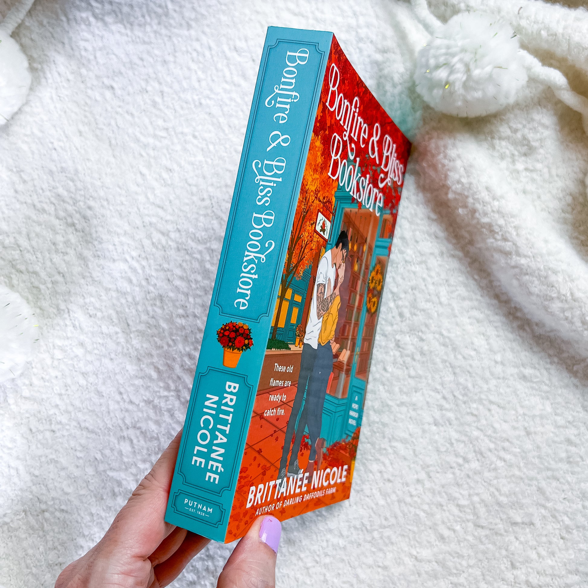 A hand holds a book titled "Bonfire & Bliss Bookstore" by Brittanie Nicole. The book has a turquoise spine and a red-orange illustrated cover, photographed against a soft white textured blanket.