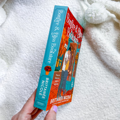 A hand holds a book titled "Bonfire & Bliss Bookstore" by Brittanie Nicole. The book has a turquoise spine and a red-orange illustrated cover, photographed against a soft white textured blanket.