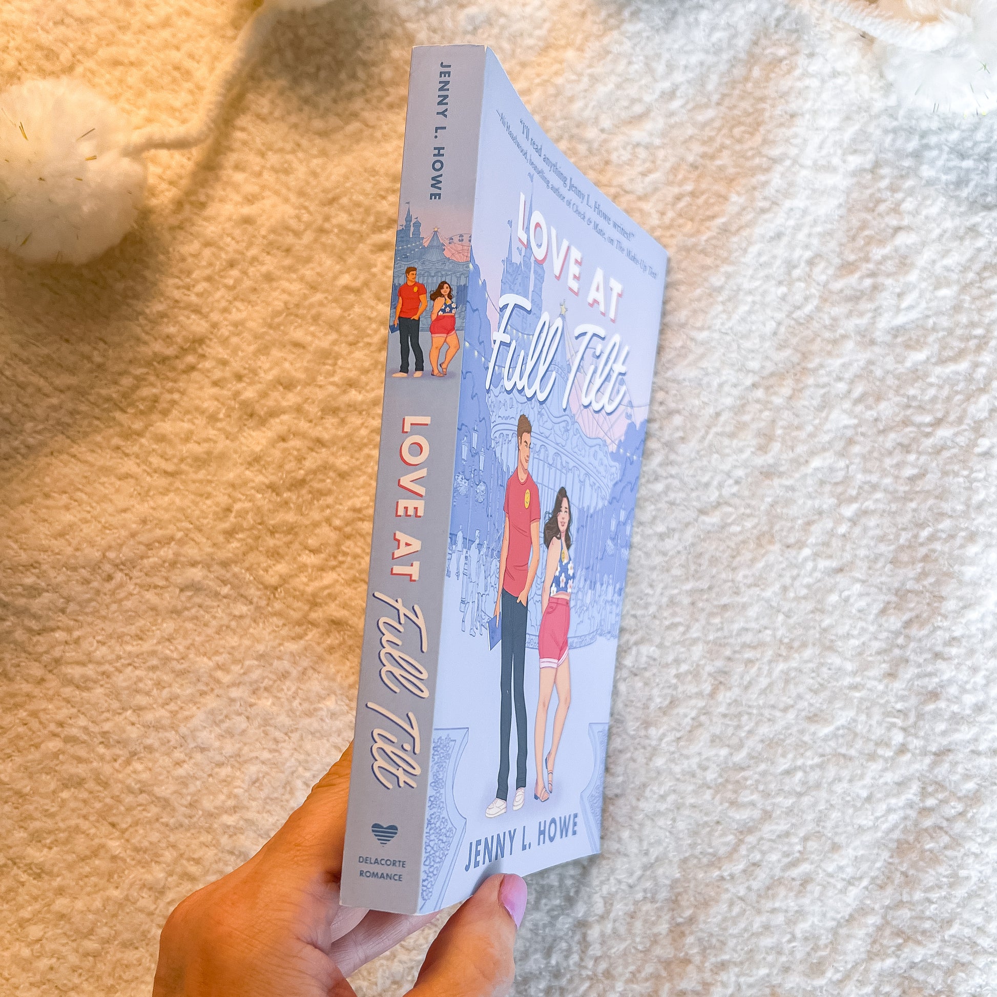 A hand holds a paperback book titled 'Love at Full Till' by Jenny L. Howe against a soft, textured cream-colored blanket background.