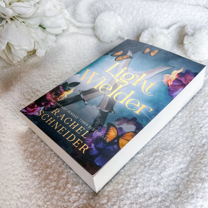 A paperback copy of the book 'Light Wielder' by Rachel Schneider, featuring a fantasy cover with swords, butterflies, and purple flowers, resting on a soft white textured blanket with white flowers in the background.