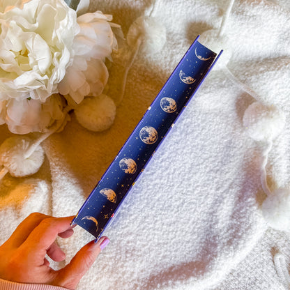 Hand holding book on edge with celestial design on a textured surface with flowers.