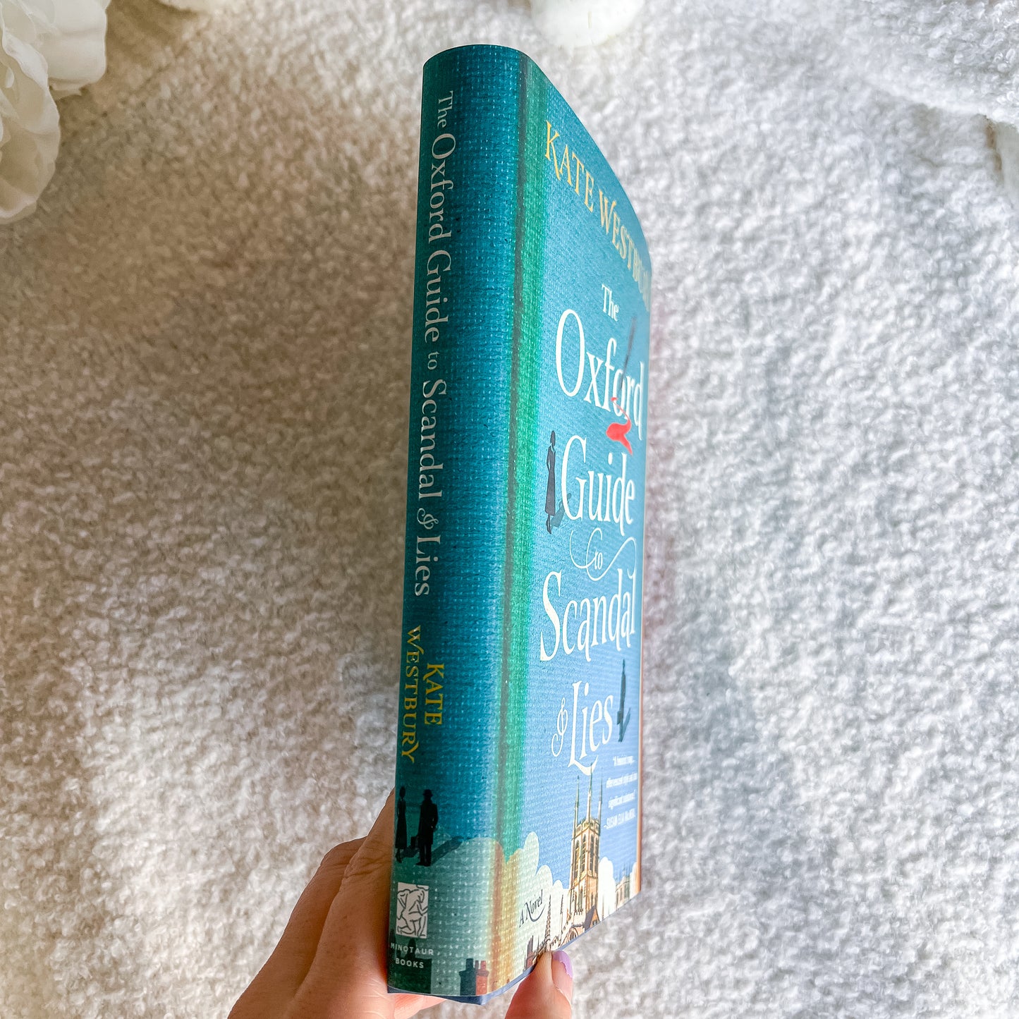 A hand holds a copy of 'The Oxford Guide to Scandal of Lies' by Kate Winslet, showing the teal-blue spine, against a soft white textured blanket background.